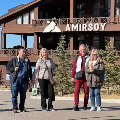 Group of four adults in winter jackets posing in front of a brown wooden lodge with a large AMIRSOY sign and pine trees.