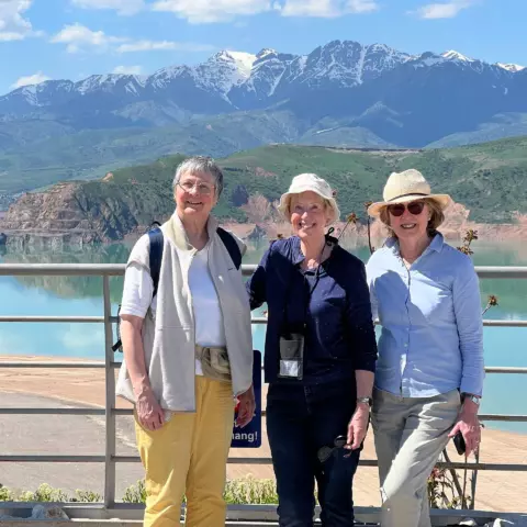 Three smiling women posing on a railing with a turquoise lake and snow‑capped mountains behind them.