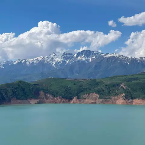 Snow-capped mountains rise above a turquoise lake under a blue sky with white clouds, landscape vista.